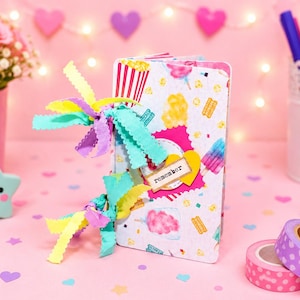 Op de afbeelding: Een decoratief notitieboek met een kleurrijk ijs- en popcornpatroon, vastgebonden met linten. Het notitieboek heeft een klein label met de tekst "remember". De achtergrond bevat roze rozen, pennen en hartvormige decoraties.