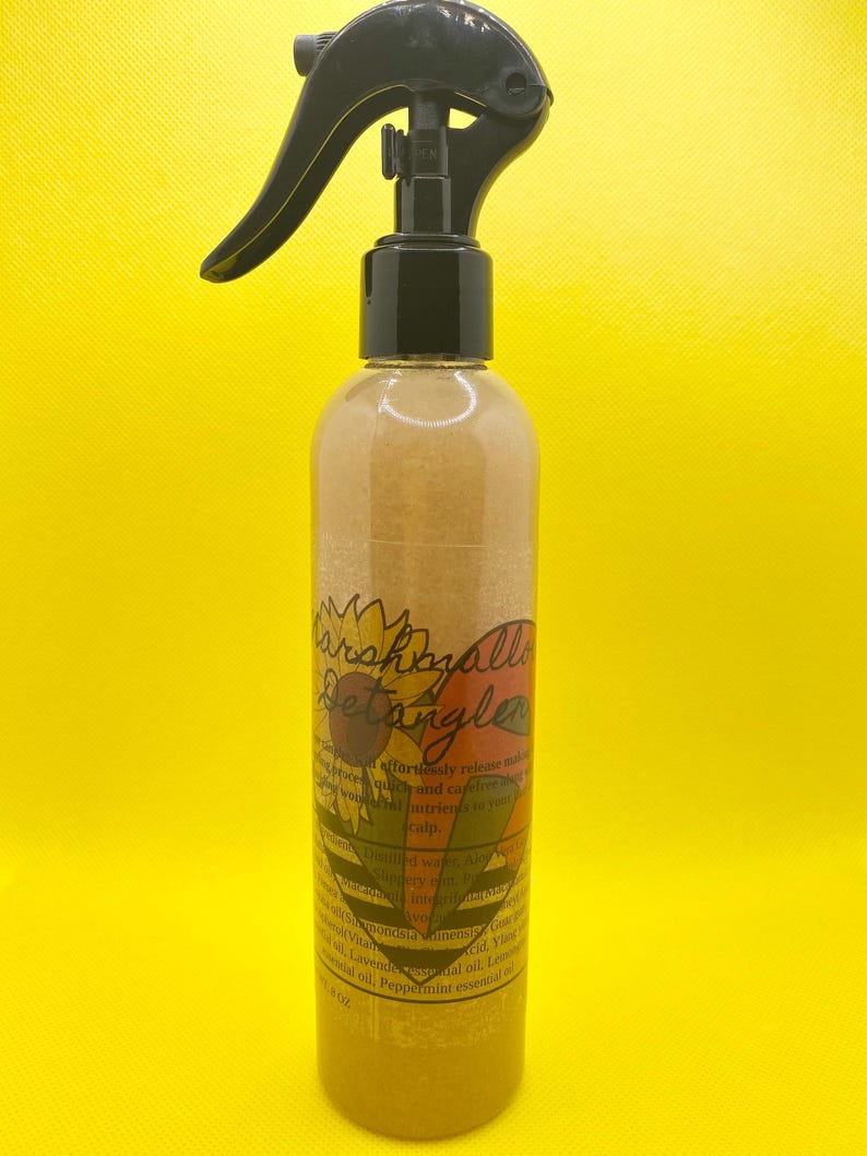 K&ouml;nnte beinhalten: Eine klare Flasche mit einer schwarzen Spr&uuml;hd&uuml;se, gef&uuml;llt mit einer goldenen Fl&uuml;ssigkeit. Die Flasche hat ein Etikett mit dem Text "Marshmallow Detangler" und einer Sonnenblumengrafik. Das Etikett listet auch die Zutaten auf, darunter Avocado&ouml;l, Lavendel&ouml;l, Zitronen&ouml;l und Pfefferminz&ouml;l.