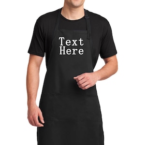 May include: A black apron with two pockets and adjustable straps. The apron has the text "Text Here" printed in white on the front.