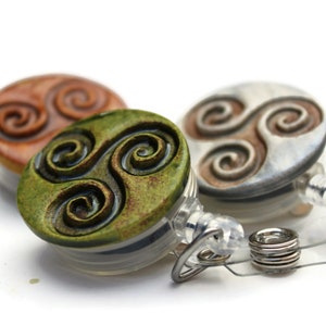 May include: Three badge reels with Celtic spiral designs. The reels are in shades of brown, green, and silver. Each reel has a clear plastic body and a retractable cord with a metal clip.