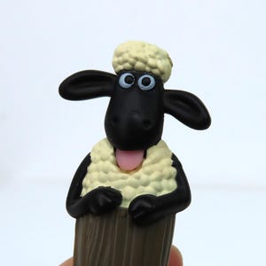 以下が含まれることがあります： ショーン・ザ・シープのフィギュア。黒い羊は、黒い顔、耳、腕を持ち、クリーム色のウールのような体と頭をしています。羊の舌はピンク色で、青い目をしています。フィギュアは白い背景にあります。