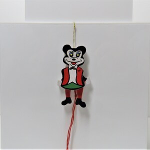 May include: A wooden pull-string toy shaped like a mouse with a red vest, green pants, and black shoes. The mouse has a black hat and a white face with black eyes and a red mouth.