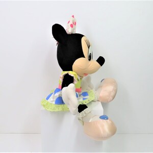 Puede incluir: Un juguete de peluche de Minnie Mouse con un vestido verde, azul y rosa con lunares blancos. Lleva un lazo rosa con lunares blancos en el pelo. Est&aacute; sentada con las piernas cruzadas y sus pies est&aacute;n en zapatillas rosas con detalles azules.