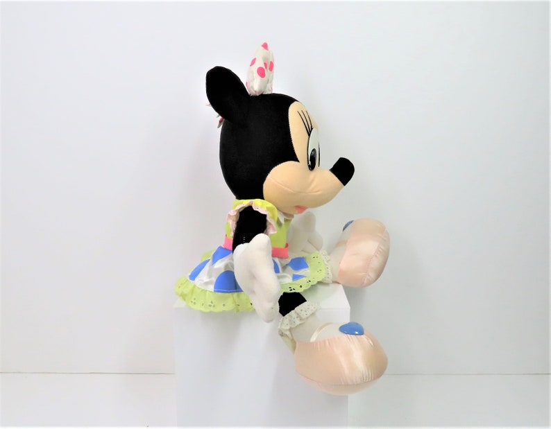 Puede incluir: Un juguete de peluche de Minnie Mouse con un vestido verde, azul y rosa con lunares blancos. Lleva un lazo rosa con lunares blancos en el pelo. Est&aacute; sentada con las piernas cruzadas y sus pies est&aacute;n en zapatillas rosas con detalles azules.
