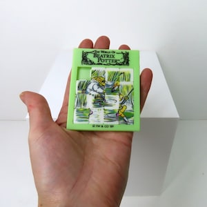 Könnte beinhalten: Ein grünes Plastikpuzzle mit einer Szene aus "Die Welt von Beatrix Potter". Das Puzzle besteht aus neun Quadraten und zeigt zwei Figuren aus dem Buch, eine angelt und die andere steht im Wasser. Das Puzzle befindet sich in einer Hand.