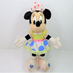 Puede incluir: Un juguete de peluche de Minnie Mouse con un vestido verde y blanco de lunares con un lazo rosa. Tiene ojos negros y una nariz negra. Lleva zapatos de ballet rosas con corazones azules.