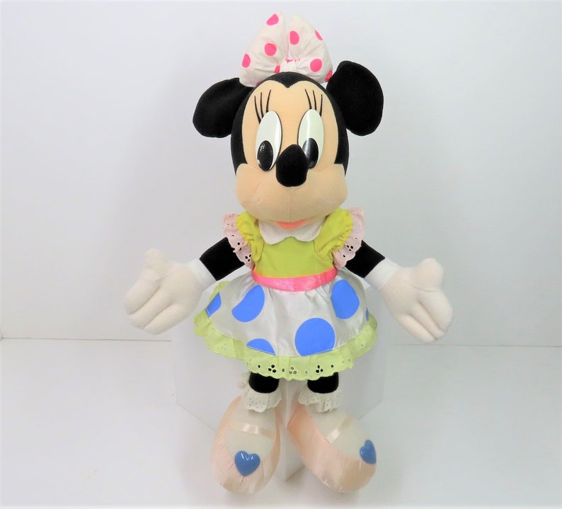 Puede incluir: Un juguete de peluche de Minnie Mouse con un vestido verde y blanco de lunares con un lazo rosa. Tiene ojos negros y una nariz negra. Lleva zapatos de ballet rosas con corazones azules.