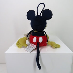 May include: A plush Mickey Mouse toy with a red and white outfit, black ears, and a black tail. The toy is sitting on a white surface and has a tag that says "GUND".