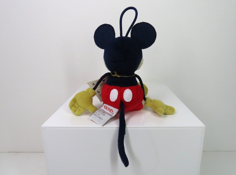 May include: A plush Mickey Mouse toy with a red and white outfit, black ears, and a black tail. The toy is sitting on a white surface and has a tag that says "GUND".