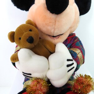 May include: A plush Mickey Mouse wearing a red, green, and gold plaid shirt, holding a brown teddy bear. The teddy bear has black eyes and a brown nose.