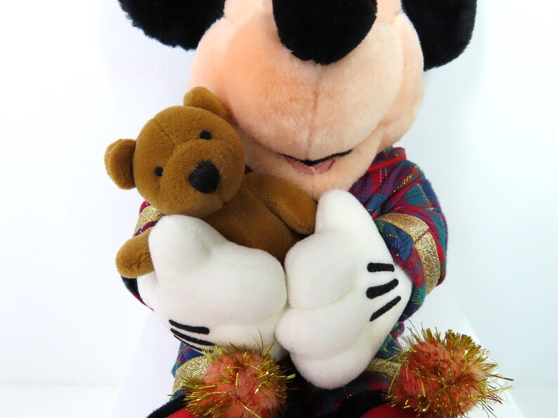May include: A plush Mickey Mouse wearing a red, green, and gold plaid shirt, holding a brown teddy bear. The teddy bear has black eyes and a brown nose.