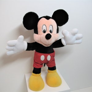 May include: A plush Mickey Mouse toy with black ears, a red and white shirt, and yellow shoes. The toy is standing on a white surface.