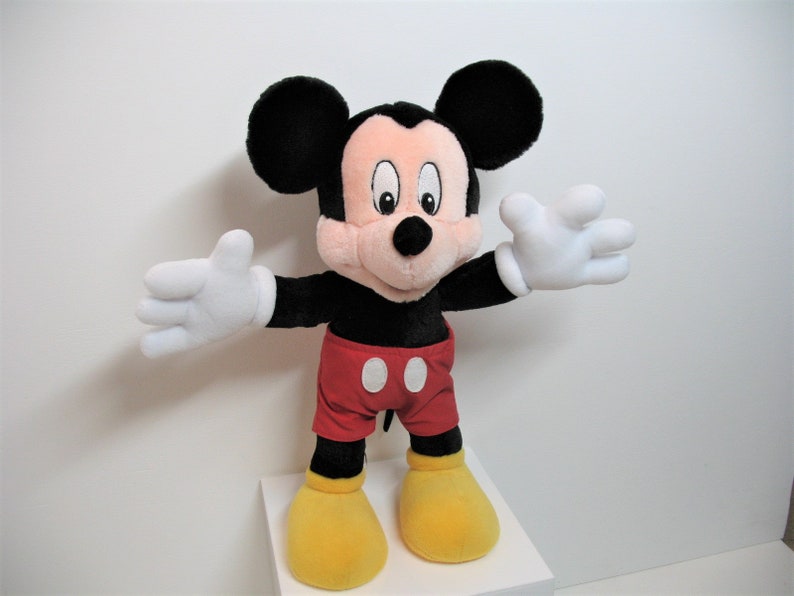 May include: A plush Mickey Mouse toy with black ears, a red and white shirt, and yellow shoes. The toy is standing on a white surface.