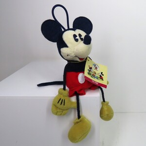 May include: A plush Mickey Mouse toy sitting on a white surface. The toy is wearing red shorts with a white circle, black shoes, and yellow gloves. The toy has a black nose and large black ears. The toy has a tag that says "GUND" on it.