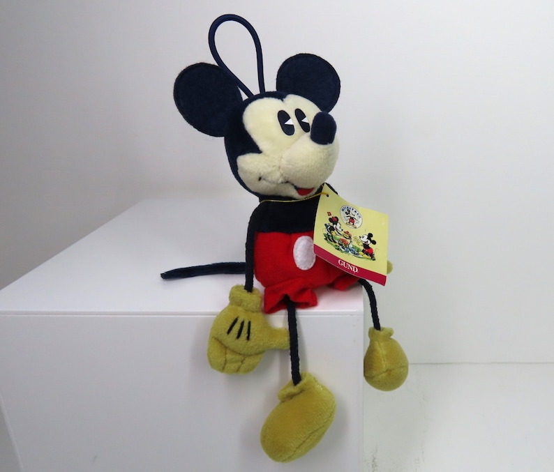 May include: A plush Mickey Mouse toy sitting on a white surface. The toy is wearing red shorts with a white circle, black shoes, and yellow gloves. The toy has a black nose and large black ears. The toy has a tag that says "GUND" on it.