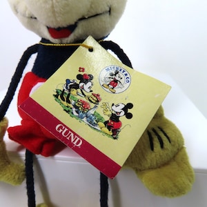 May include: A plush Mickey Mouse toy with long limbs, wearing red shorts, black shoes, and a blue shirt. The toy has a white face with black eyes and a red mouth. The toy is holding a tag that says "MICKEY & CO. GUND".