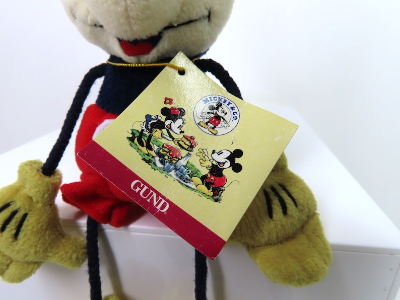 May include: A plush Mickey Mouse toy with long limbs, wearing red shorts, black shoes, and a blue shirt. The toy has a white face with black eyes and a red mouth. The toy is holding a tag that says "MICKEY & CO. GUND".
