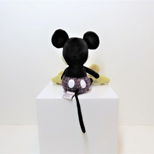 May include: A black and yellow plush Mickey Mouse toy wearing red and black plaid shorts. The toy is sitting on a white surface.