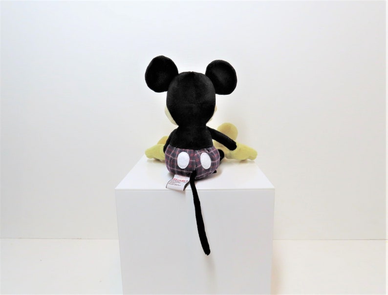 May include: A black and yellow plush Mickey Mouse toy wearing red and black plaid shorts. The toy is sitting on a white surface.