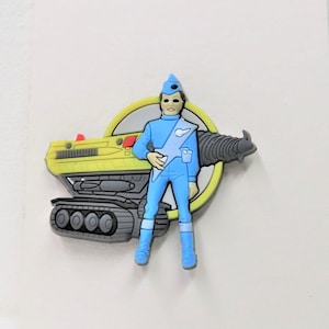 Pode incluir: Uma figura de plástico azul e amarela de um personagem do programa de televisão Thunderbirds. O personagem está de pé ao lado de um veículo amarelo e cinza com uma broca.