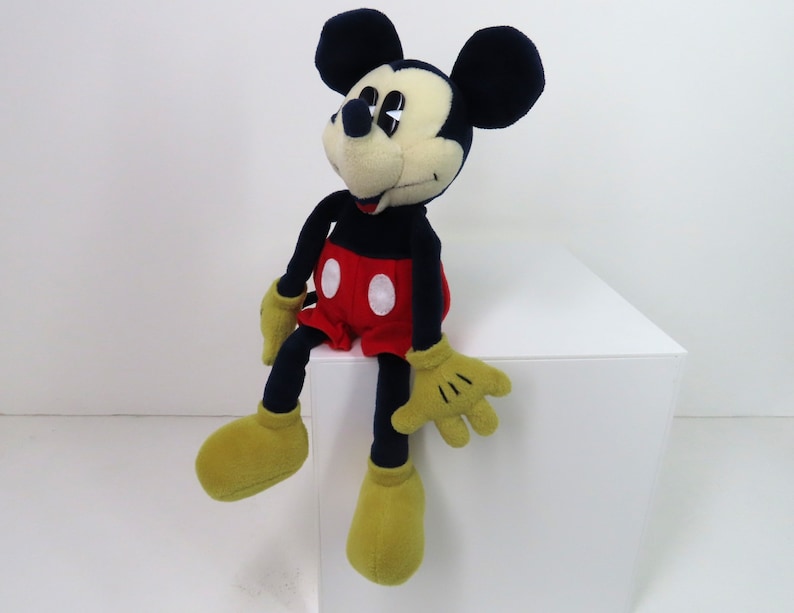 May include: A plush Mickey Mouse toy sitting on a white surface. The toy is wearing red shorts with white polka dots and yellow shoes. Mickey has black ears and a black nose.
