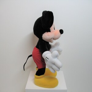 May include: A black and red plush Mickey Mouse toy with yellow shoes. The toy is standing on a white surface.