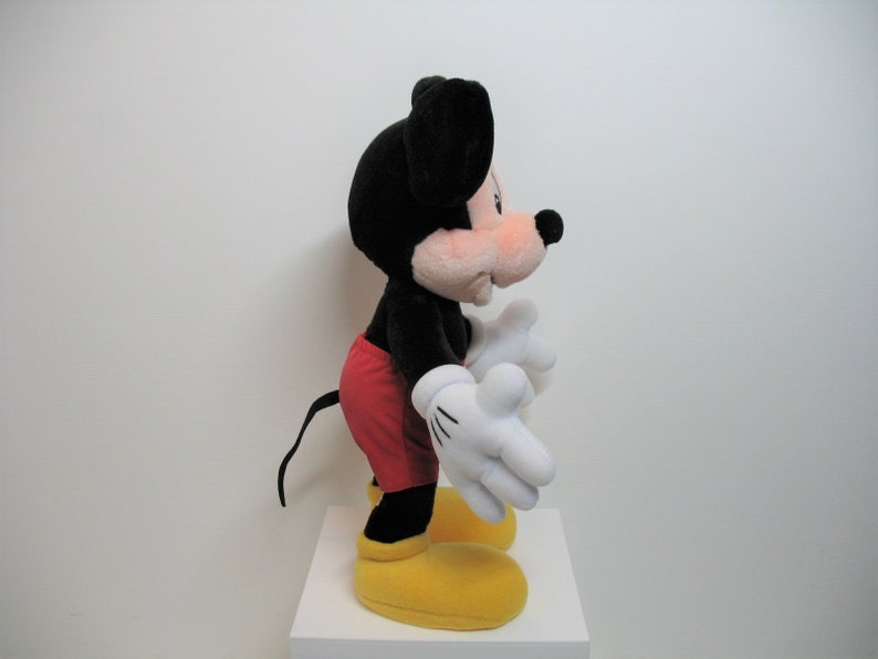 May include: A black and red plush Mickey Mouse toy with yellow shoes. The toy is standing on a white surface.