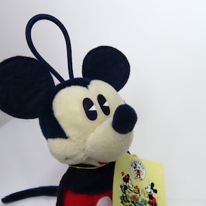 May include: A plush Mickey Mouse toy with a red and blue outfit. The toy has a loop on top of its head and a tag that says "Mickey Mouse Co."