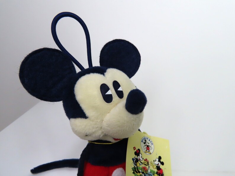 May include: A plush Mickey Mouse toy with a red and blue outfit. The toy has a loop on top of its head and a tag that says "Mickey Mouse Co."