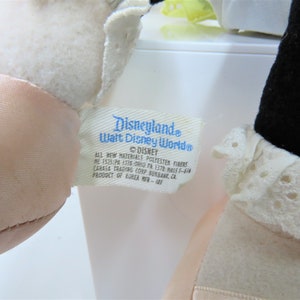 Puede incluir: Un peluche blanco con una cara rosa y un cuello de encaje blanco. El peluche tiene una etiqueta azul adjunta que dice "Disneyland Walt Disney World" y "DISNEY". La etiqueta tambi&eacute;n incluye informaci&oacute;n sobre los materiales utilizados y el fabricante.