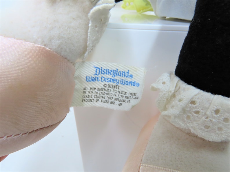 Puede incluir: Un peluche blanco con una cara rosa y un cuello de encaje blanco. El peluche tiene una etiqueta azul adjunta que dice "Disneyland Walt Disney World" y "DISNEY". La etiqueta tambi&eacute;n incluye informaci&oacute;n sobre los materiales utilizados y el fabricante.