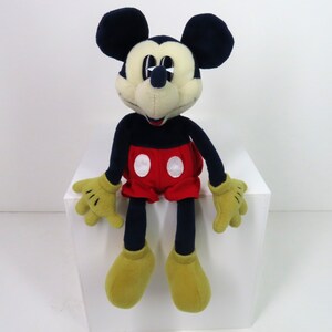 May include: A plush Mickey Mouse toy with black ears, a black body, red shorts with white circles, and yellow shoes. The toy is sitting on a white surface.