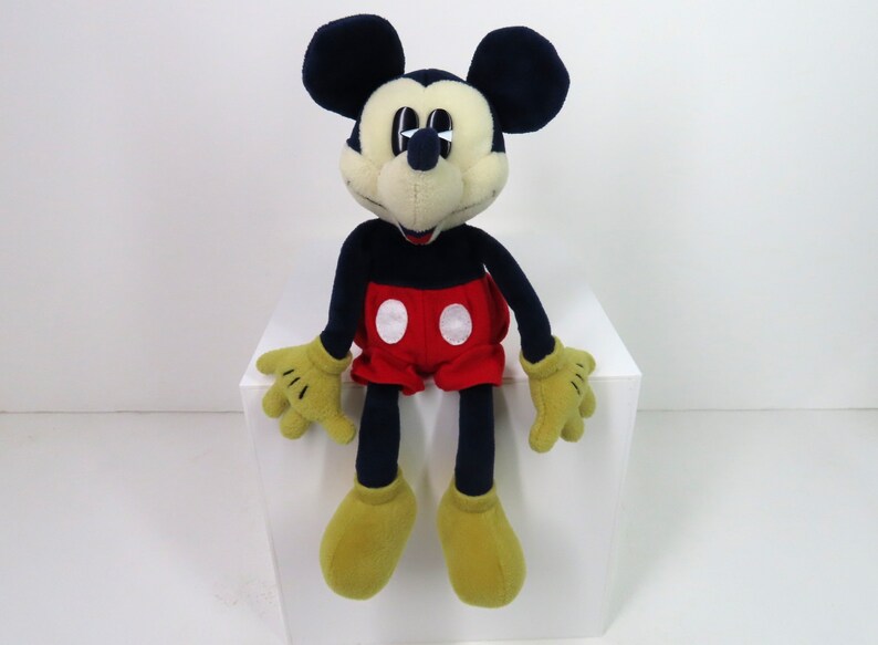 May include: A plush Mickey Mouse toy with black ears, a black body, red shorts with white circles, and yellow shoes. The toy is sitting on a white surface.