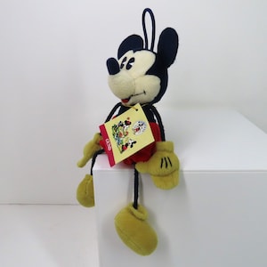 May include: A plush Mickey Mouse toy with black ears, a red and white shirt, and yellow shoes. The toy is sitting on a white surface and has a yellow tag with the word "GESSO" on it.