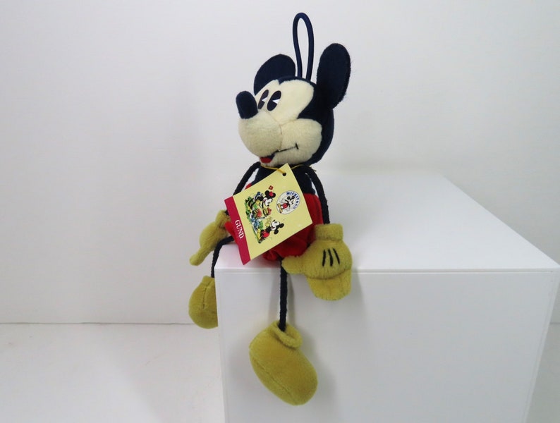 May include: A plush Mickey Mouse toy with black ears, a red and white shirt, and yellow shoes. The toy is sitting on a white surface and has a yellow tag with the word "GESSO" on it.