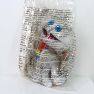 Peut inclure: Un jouet en peluche blanc avec des yeux bleus, un bec rouge et des accents colorés, emballé dans un sac en plastique transparent. Le design du jouet comprend des détails orange, jaune et bleu. Le sac est recouvert de texte imprimé.