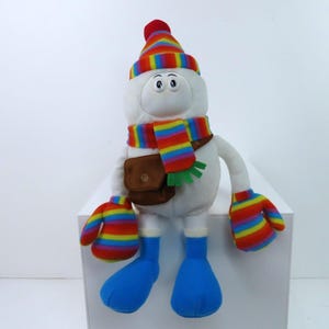 Peut inclure: Une peluche bonhomme de neige avec un corps blanc, portant un chapeau, une écharpe et des mitaines rayés arc-en-ciel. Il a une sacoche marron et des bottes bleues. Le bonhomme de neige a une expression joyeuse.