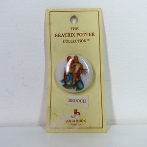 Puede incluir: Una broche de cerámica blanca con un conejo de dibujos animados con ropa azul y nariz roja. La broche está empaquetada en una tarjeta amarilla con el texto "The Beatrix Potter Collection®" y "Brooch".
