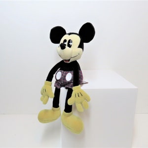May include: A black and yellow plush Mickey Mouse toy wearing red and white plaid shorts. The toy is sitting on a white surface.