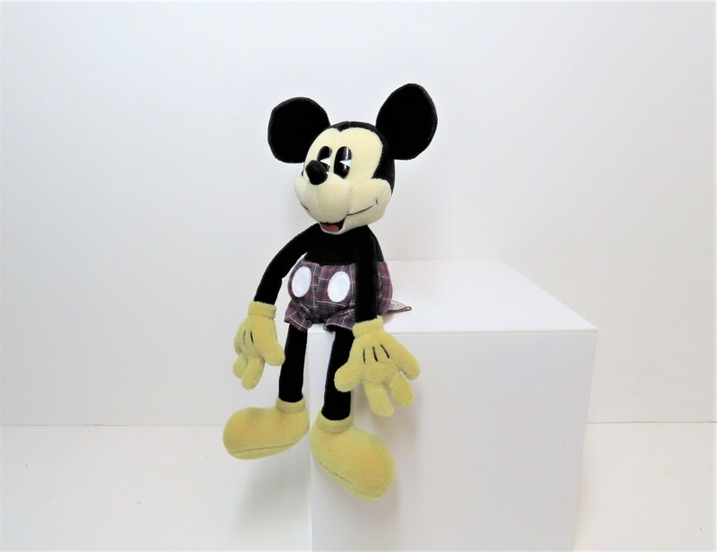 May include: A black and yellow plush Mickey Mouse toy wearing red and white plaid shorts. The toy is sitting on a white surface.