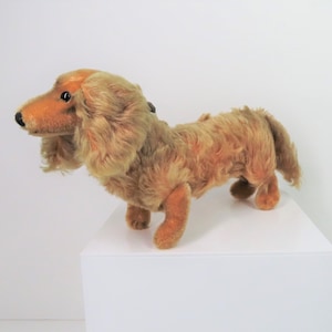 Puede incluir: Un juguete de peluche de dachshund con pelo marrón y ojos negros. El juguete está de pie sobre una superficie blanca.