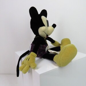 Op de afbeelding: Een zwart en geel pluche Mickey Mouse speelgoed dat een geruite kilt draagt. Het speelgoed zit op een wit oppervlak.