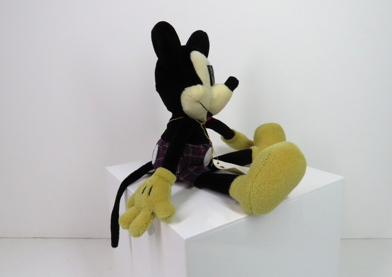 Op de afbeelding: Een zwart en geel pluche Mickey Mouse speelgoed dat een geruite kilt draagt. Het speelgoed zit op een wit oppervlak.