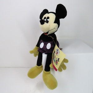 Op de afbeelding: Een zwart en geel pluche Mickey Mouse speelgoed dat een geruite rok draagt. Het speelgoed zit op een wit oppervlak en heeft een label aan zijn been.