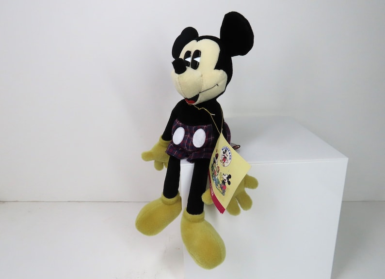 Op de afbeelding: Een zwart en geel pluche Mickey Mouse speelgoed dat een geruite rok draagt. Het speelgoed zit op een wit oppervlak en heeft een label aan zijn been.