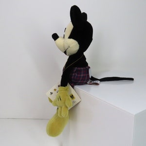Op de afbeelding: Een zwart-witte pluche Mickey Mouse die een rood-zwart geruit kilt draagt. Het pluche speelgoed zit op een wit oppervlak en heeft een geel label aan zijn been.