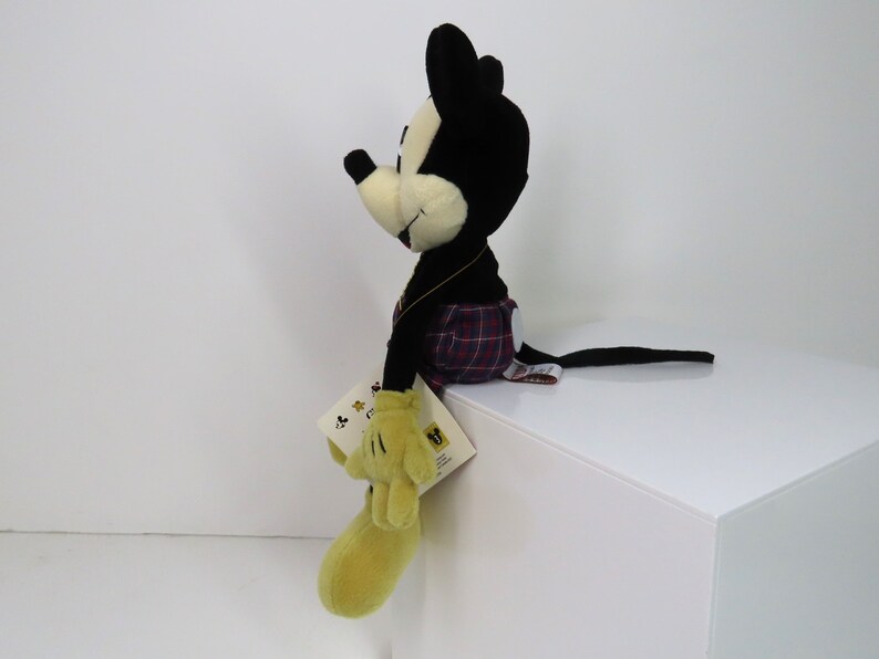Op de afbeelding: Een zwart-witte pluche Mickey Mouse die een rood-zwart geruit kilt draagt. Het pluche speelgoed zit op een wit oppervlak en heeft een geel label aan zijn been.