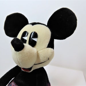 May include: A plush Mickey Mouse toy with black ears, a black nose, and large black eyes. The toy is wearing a red and black plaid kilt.