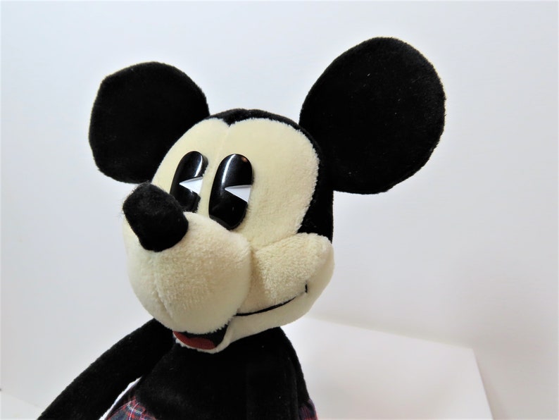 May include: A plush Mickey Mouse toy with black ears, a black nose, and large black eyes. The toy is wearing a red and black plaid kilt.