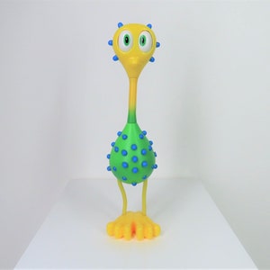 Puede incluir: Un juguete amarillo y verde con ojos azules y puntos azules por todas partes. El juguete tiene un cuello largo y dos piernas amarillas.